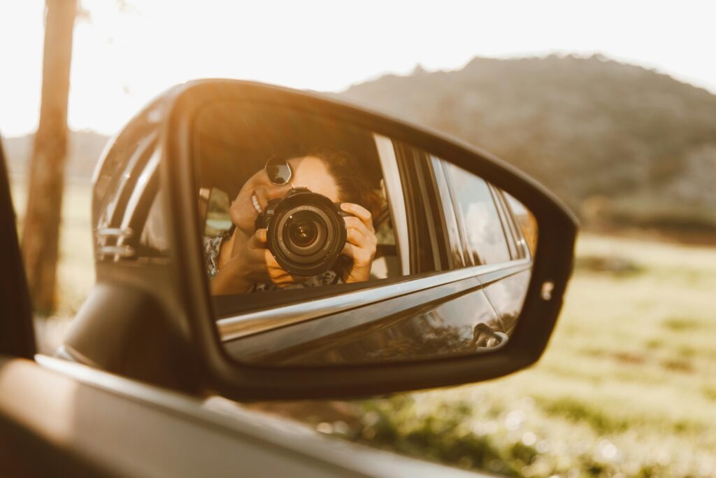 A person takes a photo through a car mirror reflection, capturing a sunny outdoor scene.